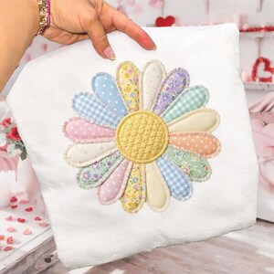 May include: White square pillow with a large, colorful daisy applique. The daisy features pastel petals in various patterns, including polka dots, gingham, and floral prints, surrounding a textured yellow center. The pillow is held up by a hand.