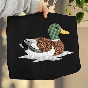 Peut inclure: Sac fourre-tout noir avec une application de canard. Le canard a une tête et un cou à carreaux verts et bruns, un bec jaune et un corps à motifs floraux bruns. Les ailes sont grises et blanches, et le canard semble flotter sur l'eau. Design fait main.