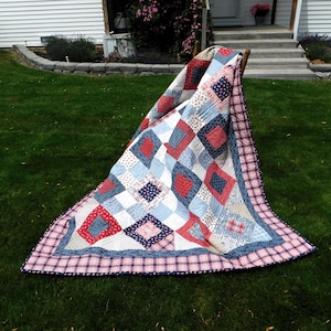 May include: A red, white, and blue patchwork quilt with a plaid border. The quilt is folded and draped over a wooden chair on a grassy lawn.