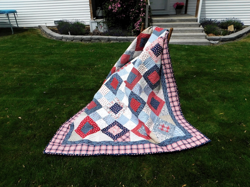 May include: A red, white, and blue patchwork quilt with a plaid border. The quilt is folded and draped over a wooden chair on a grassy lawn.