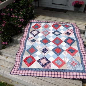 May include: A red, white, and blue patchwork quilt with a diamond pattern. The quilt is made of various fabrics, including plaid, gingham, and floral prints. It is lying on a wooden porch.