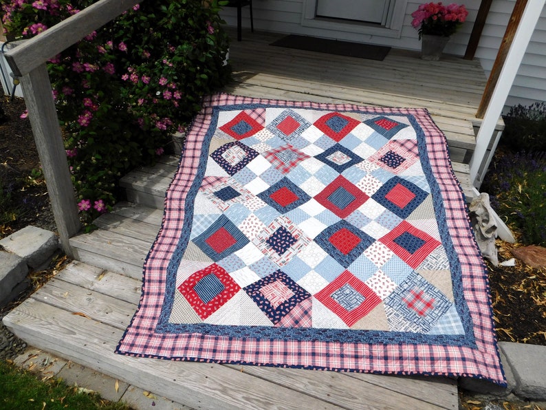 May include: A red, white, and blue patchwork quilt with a diamond pattern. The quilt is made of various fabrics, including plaid, gingham, and floral prints. It is lying on a wooden porch.