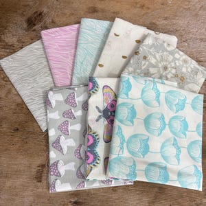 May include: A collection of folded fabric squares with various patterns. Designs include mushrooms, butterflies, and floral motifs in colors like gray, pink, and teal. The fabrics are arranged on a wooden surface.