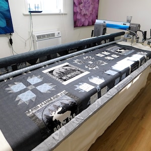 May include: A longarm quilting machine with a grey and white quilt on the bed. The quilt features a variety of designs, including a moose, trees, and the words "Roam Far" and "Memories".
