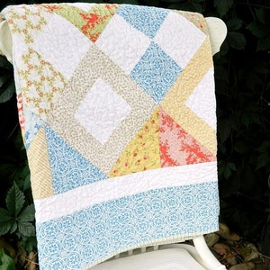May include: A white, blue, yellow, and pink patchwork quilt with a diamond pattern. The quilt is folded and draped over a white chair.