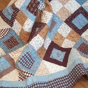 May include: A blue, brown, and white patchwork quilt with a diamond pattern. The quilt is folded in half and is lying on a wooden floor.