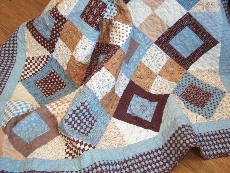May include: A blue, brown, and white patchwork quilt with a diamond pattern. The quilt is folded in half and is lying on a wooden floor.
