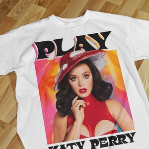 May include: White t-shirt with a graphic of Katy Perry. The word "PLAY" is above her image, and "KATY PERRY" is below. The graphic shows Katy Perry in a red outfit and a mushroom-shaped hat.