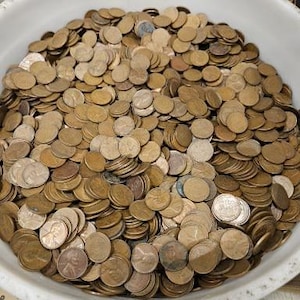 May include: A white bowl filled with a large quantity of copper-colored US pennies. The pennies are stacked in piles and are arranged in a random pattern.