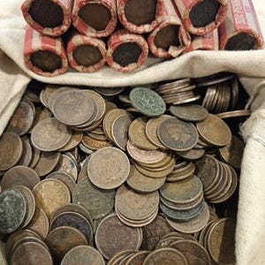 May include: A white canvas bag filled with a large collection of copper-colored coins. The bag is sitting on top of a pile of rolled up red and white paper tubes.