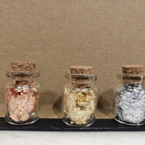May include: Three small glass bottles with cork tops, each containing a different type of metallic leaf. The bottles are arranged in a row from left to right: copper, gold, and silver.