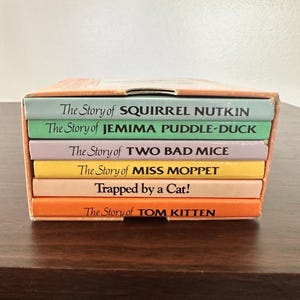 May include: A collection of children's books in a box, featuring titles such as "The Story of Squirrel Nutkin" and "The Story of Jemima Puddle-Duck." The spines are in various colours, and the books are neatly stacked in a cardboard box.