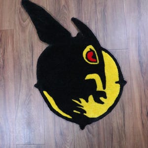 May include: A black and yellow rug in the shape of a stylized rabbit head. The rug has a black body with yellow accents and a red eye. The rug is placed on a wooden floor.