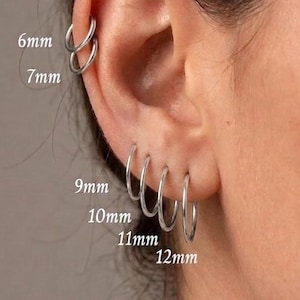 Puede incluir: Un conjunto de seis pendientes de aro plateados en una oreja, etiquetados con las medidas 6mm, 7mm, 9mm, 10mm, 11mm y 12mm. Los dos aros más pequeños están en el cartílago superior, mientras que los cuatro más grandes están en el lóbulo.