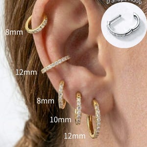 May include: A collection of gold-toned hoop earrings and a straight bar earring worn on an ear. The jewelry features clear stones and is labeled with sizes 8mm, 10mm, and 12mm. An inset shows a silver-toned D Shape Hoop with a hinged mechanism.