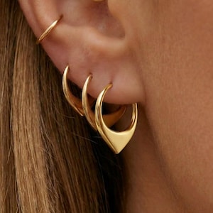 Könnte beinhalten: Ein Set aus goldfarbenen Ohrringen, bestehend aus einem kleinen schlichten Ring oben, einem mittleren Ring und einem größeren Ring mit einer markanten, spitz zulaufenden Tropfenform am unteren Rand. Der Schmuck hat eine glatte, polierte Oberfläche.