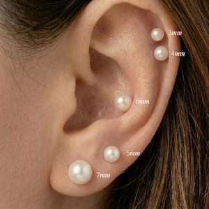 May include: A set of five round white pearl stud earrings worn in a single ear. The pearls are labelled with their respective sizes: 3mm, 4mm, 5mm, 6mm, and 7mm. Each pearl has a smooth, lustrous finish and is positioned along the ear cartilage and lobe.