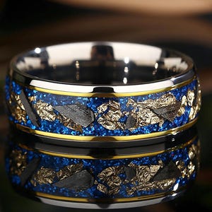 May include: A silver ring with a blue and gold inlay. The inlay features small pieces of gold and black metal, resembling a galaxy.