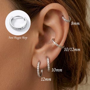 Może przedstawiać: Zestaw srebrzystych kolczyków typu huggie z cyrkoniami. Obraz przedstawia różne rozmiary oznaczone jako 8mm, 10mm, 12mm i 10/12mm, pokazując kolczyki noszone w różnych przekłuciach ucha wraz z okrągłym detalem w powiększeniu.