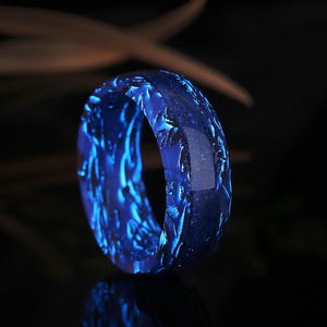 Op de afbeelding: Een gloeiende blauwe ring met een getextureerd oppervlak. De ring heeft een donkere achtergrond en de blauwe kleur is levendig en opvallend. Het ontwerp van de ring bevat ingewikkelde patronen, wat zorgt voor een unieke en artistieke uitstraling.
