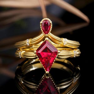 Könnte beinhalten: Ein goldener Ring mit zwei roten Rubin-Edelsteinen. Der größere Rubin ist quadratisch geschliffen, der kleinere hat eine Birnenform. Der Ring ist mit kleinen Diamanten verziert.