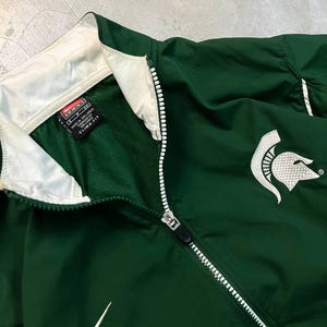 May include: Green Nike athletic jacket with a white zipper and collar. Features a white Nike swoosh logo and a Spartan helmet emblem. The jacket has a Clima-Fit label.