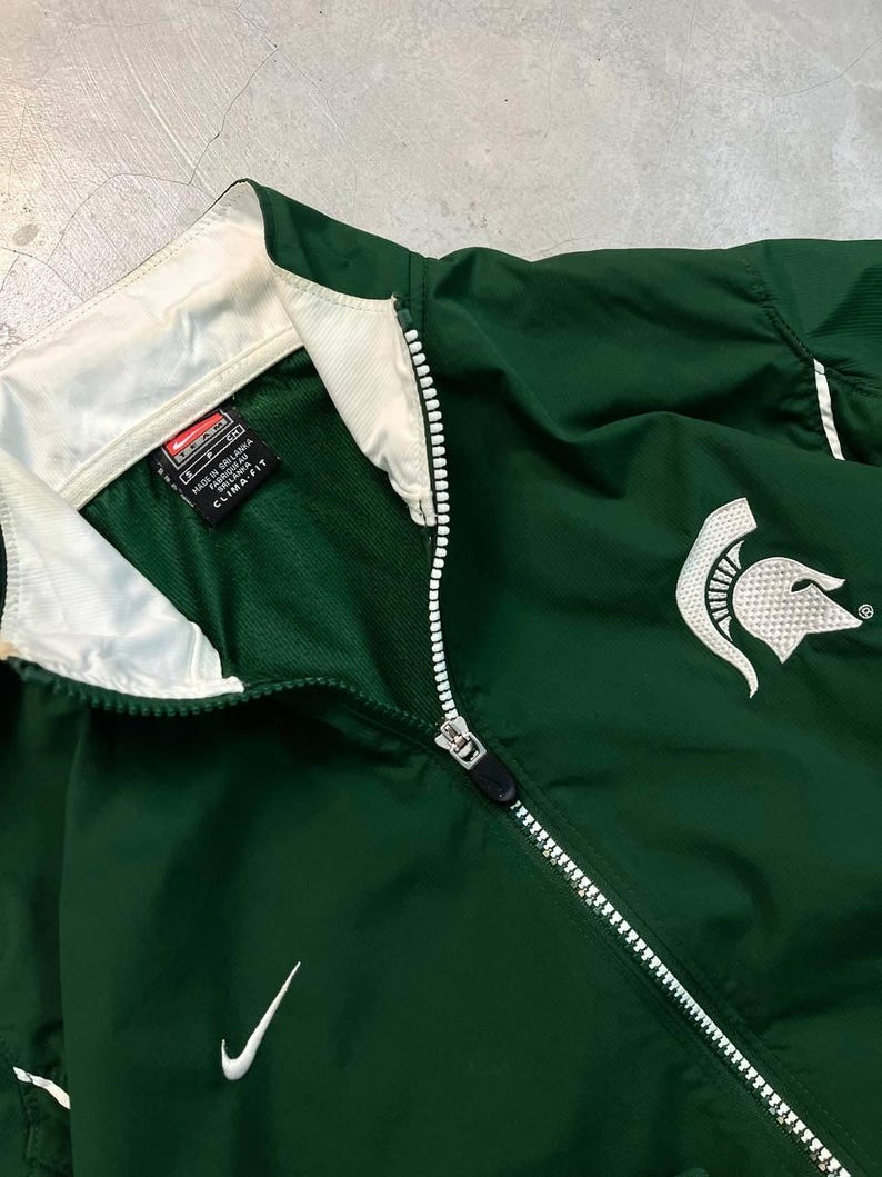May include: Green Nike athletic jacket with a white zipper and collar. Features a white Nike swoosh logo and a Spartan helmet emblem. The jacket has a Clima-Fit label.