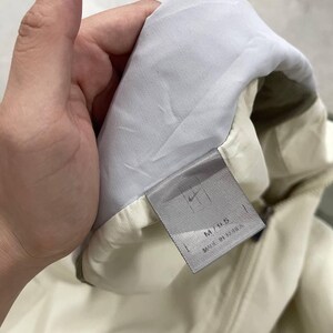 Peut inclure: Veste cr&egrave;me avec une doublure blanche et une &eacute;tiquette grise. L'&eacute;tiquette porte le logo Nike et le texte "M/95 MADE IN KOREA". La veste est tenue par une main.