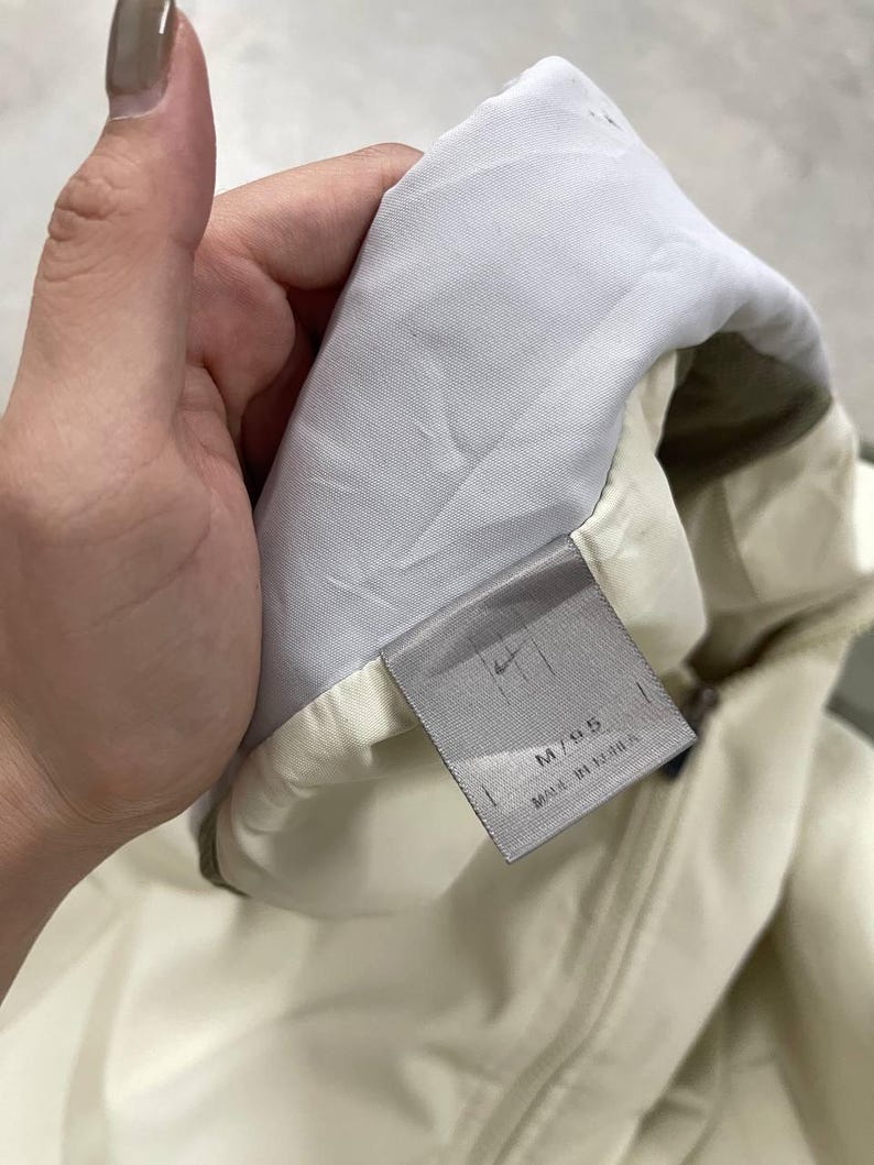 Peut inclure: Veste cr&egrave;me avec une doublure blanche et une &eacute;tiquette grise. L'&eacute;tiquette porte le logo Nike et le texte "M/95 MADE IN KOREA". La veste est tenue par une main.