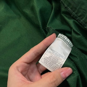 May include: Close-up of a dark green fabric with a tag attached. The tag is white with black text, including the words "100% POLYESTER" and "MADE IN SRI LANKA". The fabric appears to be a soft, textured material.