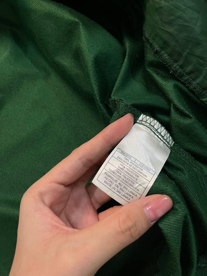 May include: Close-up of a dark green fabric with a tag attached. The tag is white with black text, including the words "100% POLYESTER" and "MADE IN SRI LANKA". The fabric appears to be a soft, textured material.