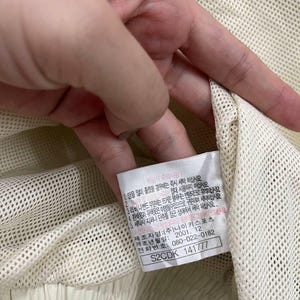 Peut inclure: Gros plan sur un v&ecirc;tement en tissu maill&eacute; cr&egrave;me avec une &eacute;tiquette. L'&eacute;tiquette contient du texte en cor&eacute;en, ainsi qu'un code produit et un num&eacute;ro de t&eacute;l&eacute;phone. Le v&ecirc;tement semble faire partie d'une tenue de sport.