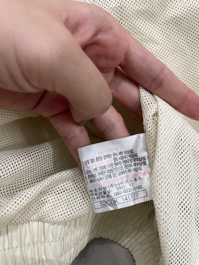 Peut inclure: Gros plan sur un v&ecirc;tement en tissu maill&eacute; cr&egrave;me avec une &eacute;tiquette. L'&eacute;tiquette contient du texte en cor&eacute;en, ainsi qu'un code produit et un num&eacute;ro de t&eacute;l&eacute;phone. Le v&ecirc;tement semble faire partie d'une tenue de sport.