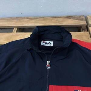 May include: A navy blue windbreaker jacket featuring a high collar, a full-length front zipper with a branded Fila pull tab, and a white interior label with the text Fila Biella-Italia. The jacket includes red color-blocked panels on the sleeves.