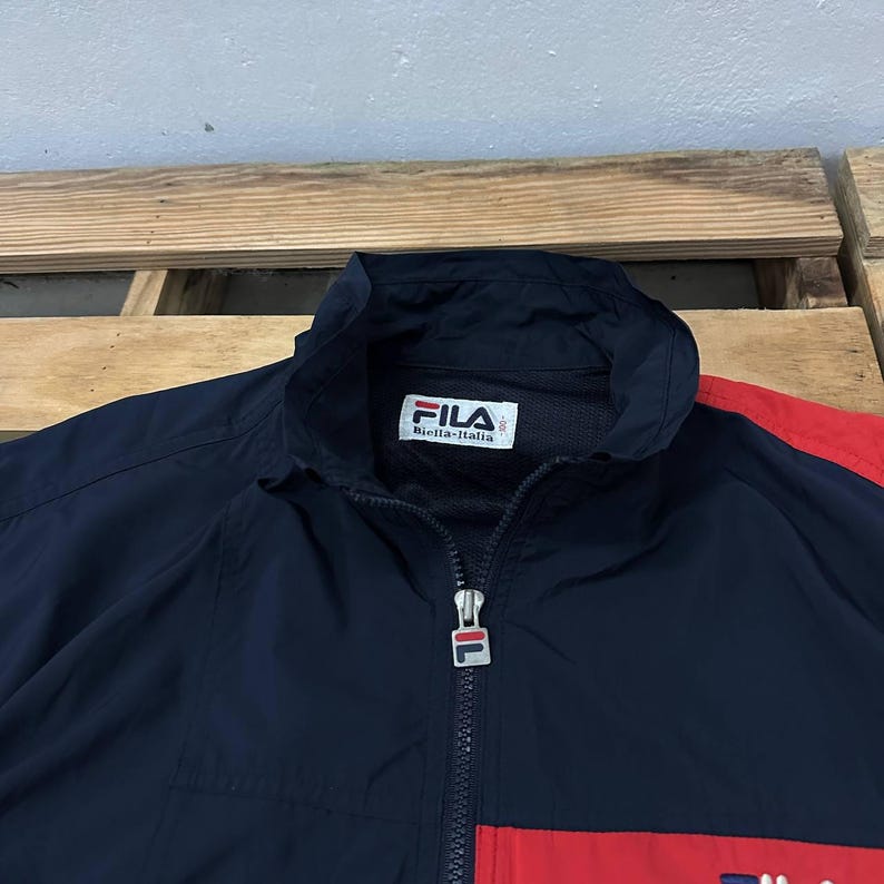 May include: A navy blue windbreaker jacket featuring a high collar, a full-length front zipper with a branded Fila pull tab, and a white interior label with the text Fila Biella-Italia. The jacket includes red color-blocked panels on the sleeves.