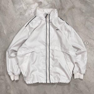 May include: A white windbreaker jacket featuring a full-length front zipper with black trim, a high collar, and long sleeves with ribbed cuffs. A small black Nike swoosh logo is embroidered on the upper left chest area of the garment.