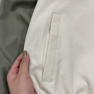 Peut inclure: Gros plan sur une section de veste. La veste est principalement blanc cass&eacute; avec un d&eacute;tail de poche rectangulaire. Une section de tissu vert olive est visible &agrave; gauche. La veste semble &ecirc;tre faite d'une mati&egrave;re lisse, peut-&ecirc;tre synth&eacute;tique.