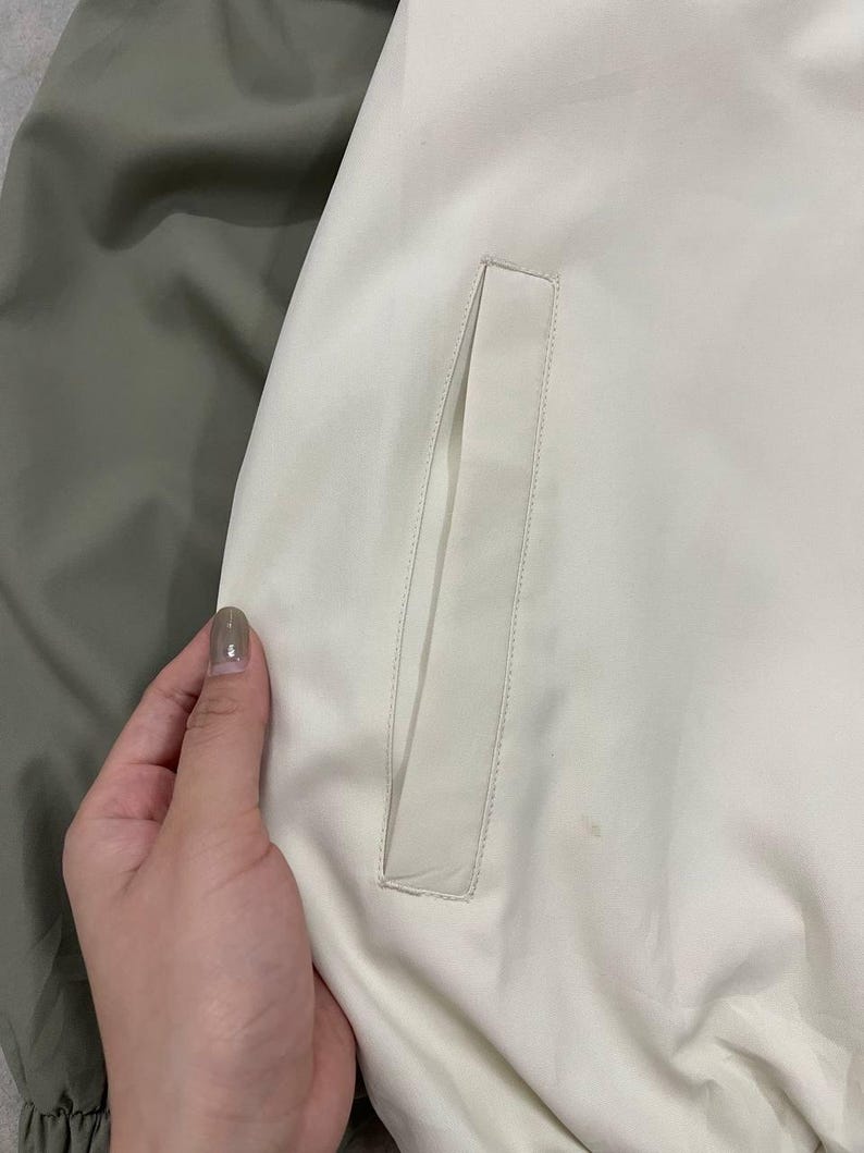 Peut inclure: Gros plan sur une section de veste. La veste est principalement blanc cass&eacute; avec un d&eacute;tail de poche rectangulaire. Une section de tissu vert olive est visible &agrave; gauche. La veste semble &ecirc;tre faite d'une mati&egrave;re lisse, peut-&ecirc;tre synth&eacute;tique.