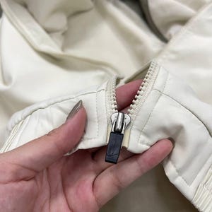 Peut inclure: Gros plan sur une veste cr&egrave;me avec une fermeture &eacute;clair argent&eacute;e et une tirette noire. La veste semble &ecirc;tre faite d'un mat&eacute;riau l&eacute;ger, avec un motif matelass&eacute; sur le devant.