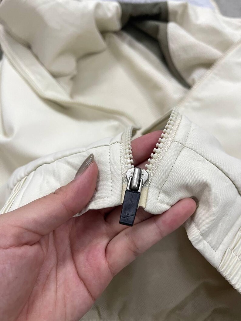 Peut inclure: Gros plan sur une veste cr&egrave;me avec une fermeture &eacute;clair argent&eacute;e et une tirette noire. La veste semble &ecirc;tre faite d'un mat&eacute;riau l&eacute;ger, avec un motif matelass&eacute; sur le devant.