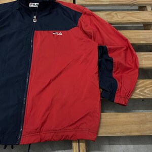 May include: A color-blocked windbreaker jacket featuring a navy blue and red design with a full-length front zipper. The jacket displays the white Fila logo on the upper left chest and a branded zipper pull. It has long sleeves with elastic cuffs.