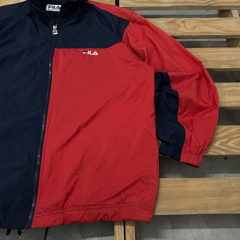 May include: A color-blocked windbreaker jacket featuring a navy blue and red design with a full-length front zipper. The jacket displays the white Fila logo on the upper left chest and a branded zipper pull. It has long sleeves with elastic cuffs.