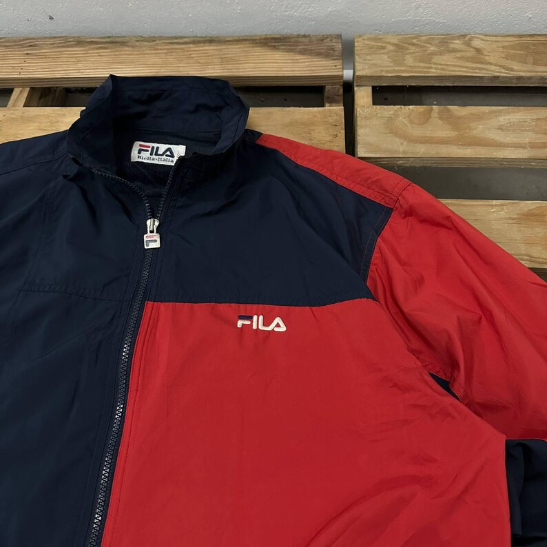 May include: A navy blue and red color-blocked windbreaker jacket featuring a high collar, a full-length front zipper with a branded pull tab, and the FILA logo embroidered in white on the chest. The interior neck label also displays the FILA brand.