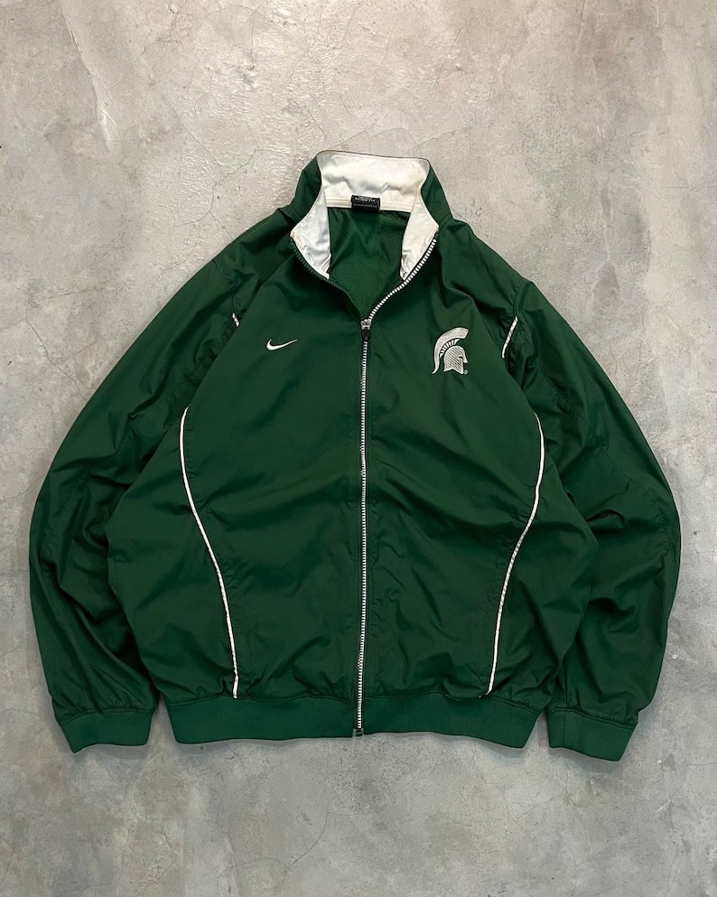 May include: A green zip-up jacket with white trim and a white collar. The jacket features a Nike logo on the chest and a Spartan helmet logo. The jacket is made of a lightweight material.
