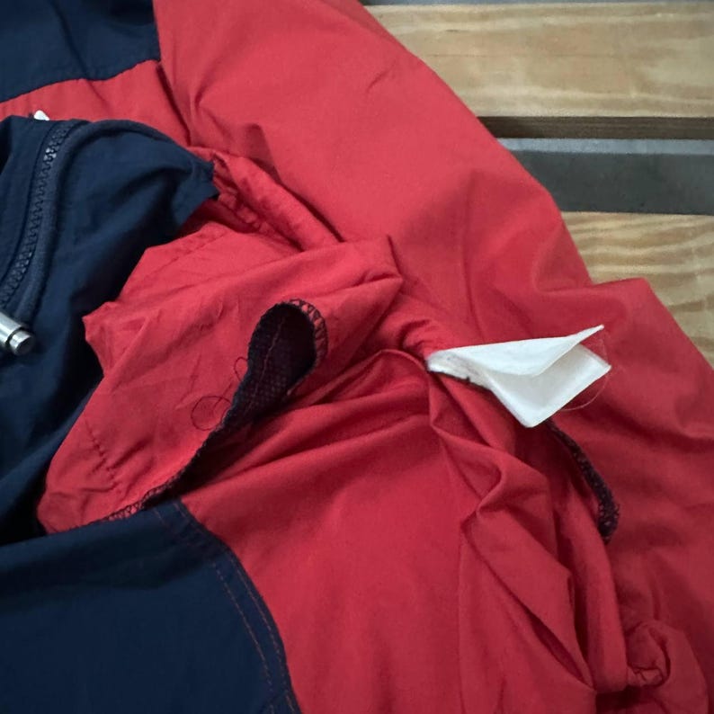 May include: A jacket featuring a color-blocked design with bold red and dark navy blue panels. The garment includes a dark navy blue zipper closure, visible stitching along the seams, and a white fabric care label attached to the interior lining.