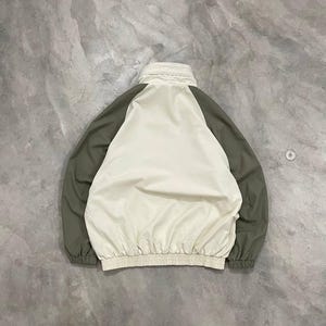 Peut inclure: Veste cr&egrave;me avec des manches vert olive. La veste a un col montant et une taille fronc&eacute;e. Le dos de la veste est visible, mettant en valeur le design en blocs de couleurs. La veste est faite d'un mat&eacute;riau l&eacute;ger.