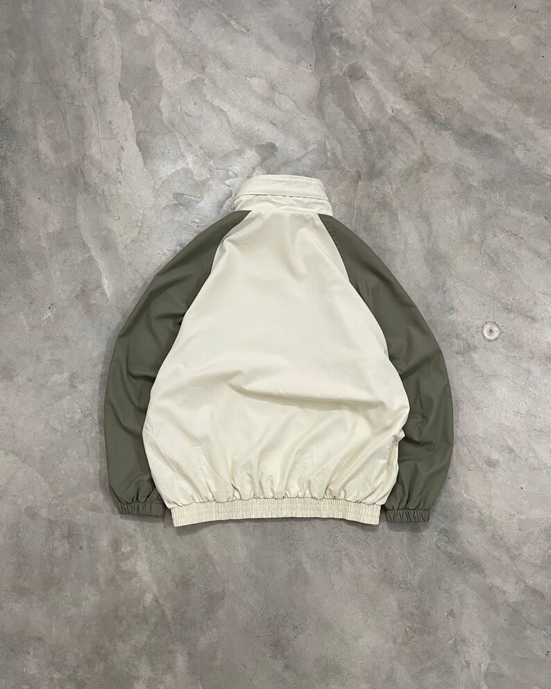 Peut inclure: Veste cr&egrave;me avec des manches vert olive. La veste a un col montant et une taille fronc&eacute;e. Le dos de la veste est visible, mettant en valeur le design en blocs de couleurs. La veste est faite d'un mat&eacute;riau l&eacute;ger.