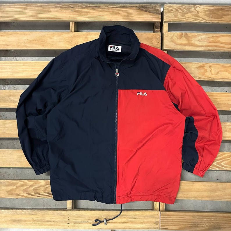 May include: A navy blue and red color-blocked windbreaker jacket featuring a full-length front zipper with a branded pull tab. The jacket has a high collar, elastic cuffs, and the FILA logo embroidered in white on the chest and inner neck label.