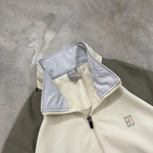 Peut inclure: Veste zipp&eacute;e cr&egrave;me avec col bleu clair et manches vert olive. La veste pr&eacute;sente un petit logo sur la poitrine et une fermeture &eacute;clair compl&egrave;te. La veste est faite d'un mat&eacute;riau l&eacute;ger.