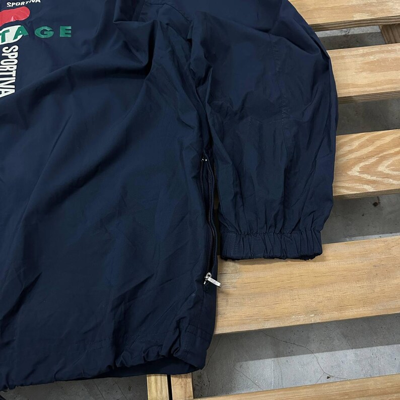 May include: A dark navy blue long-sleeve windbreaker jacket featuring a side zipper closure and elastic cuffs. The front displays partial white and green text that reads SPORTIVA and VINTAGE, along with a red graphic element near the top neckline.